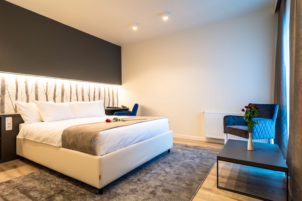 A modern bedroom with a large bed, bedside tables, a nightstand, a small table with a vase, and a chair.