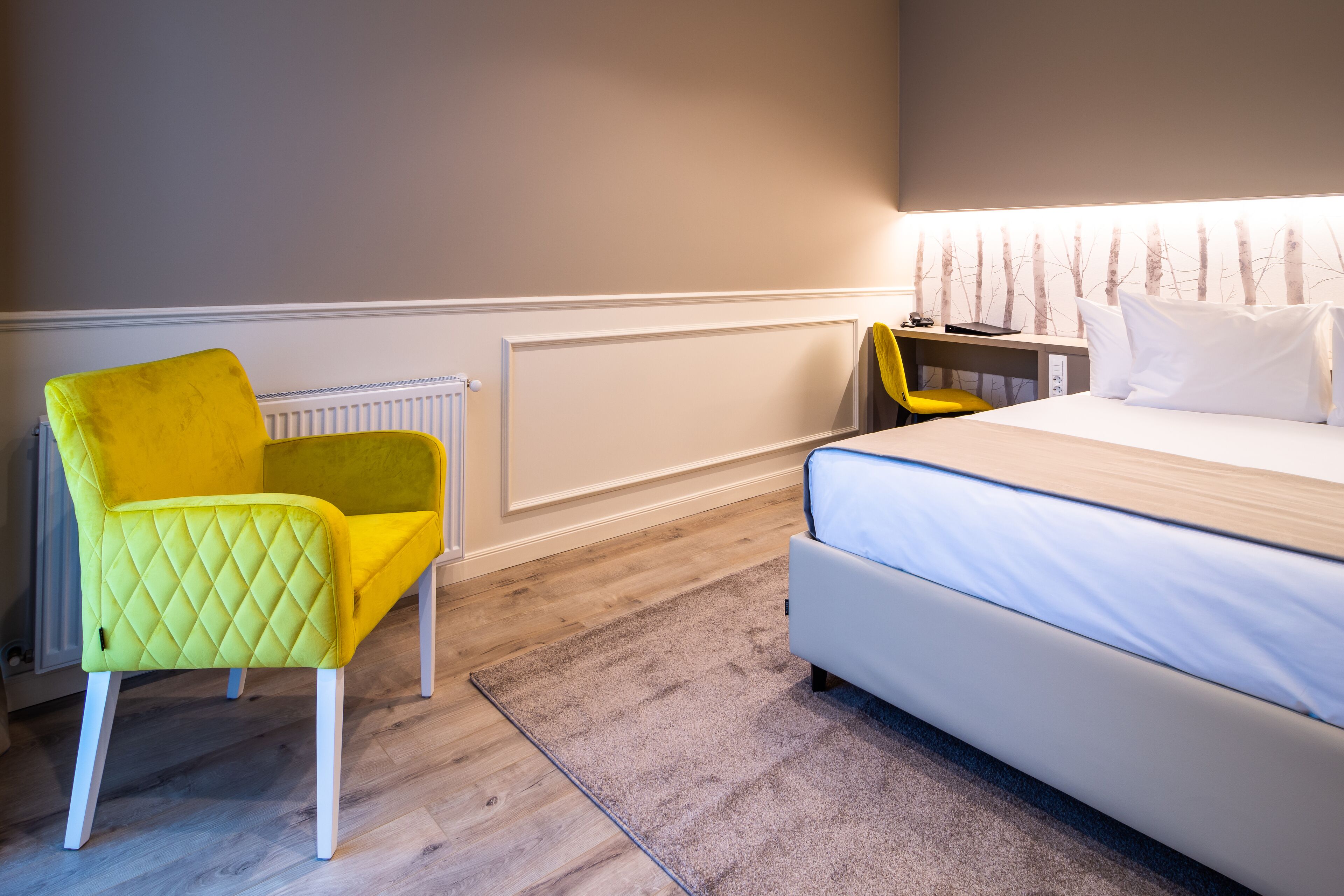 A hotel room with a bed, a yellow chair, a desk, and a radiator.