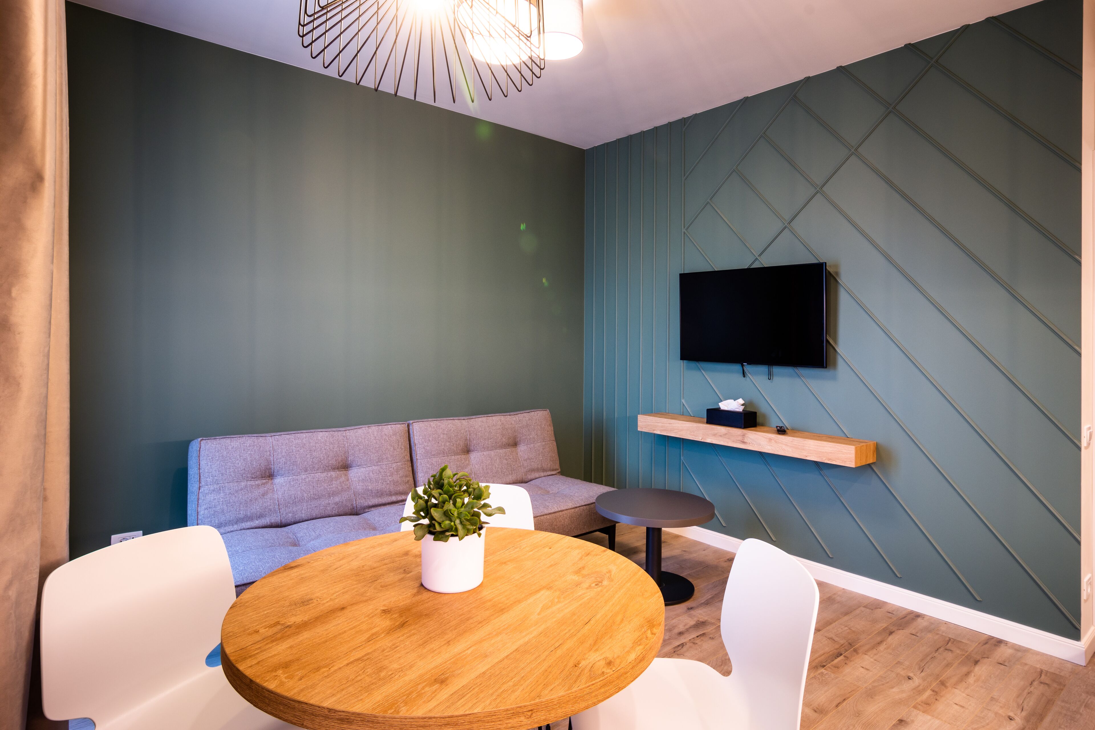 Een moderne woonkamer met een grijze bank, een ronde houten tafel met een plant in pot, en een aan de muur bevestigde televisie.