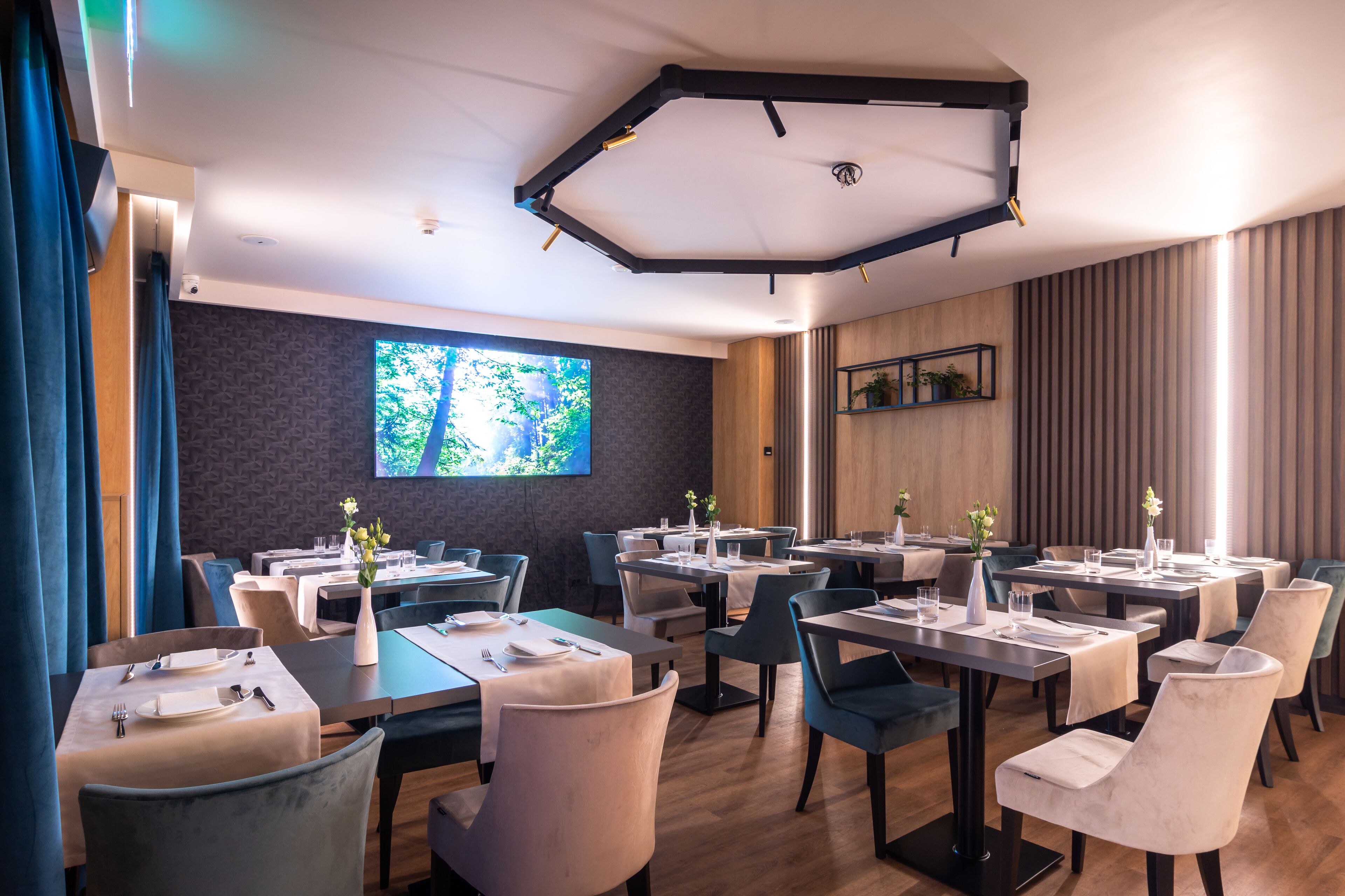 A modern dining area with multiple tables and chairs, a large screen on the wall, and a wooden accent wall.