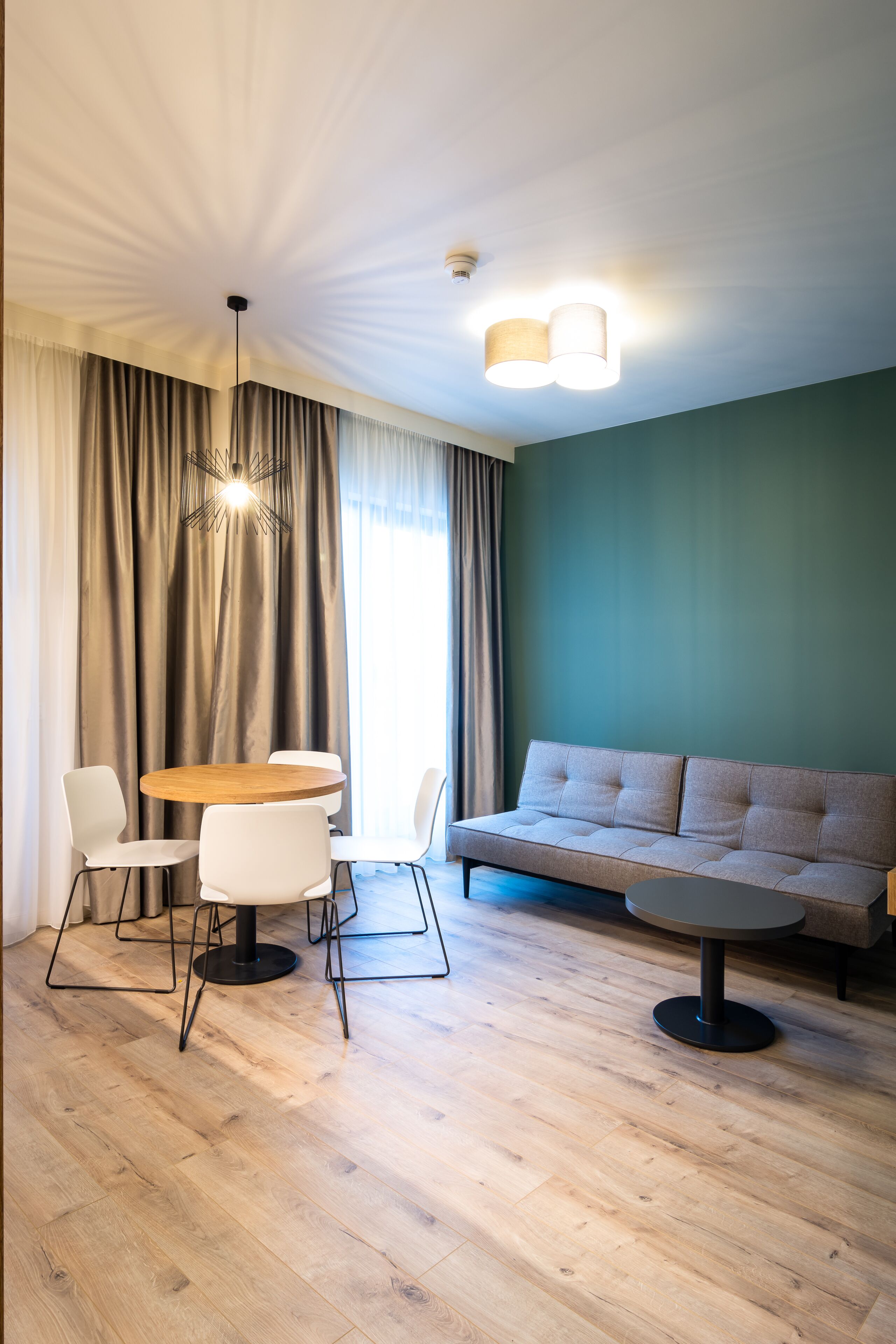 A modern living room with a wooden floor, a round dining table with four chairs, a sofa, and a ceiling light fixture.