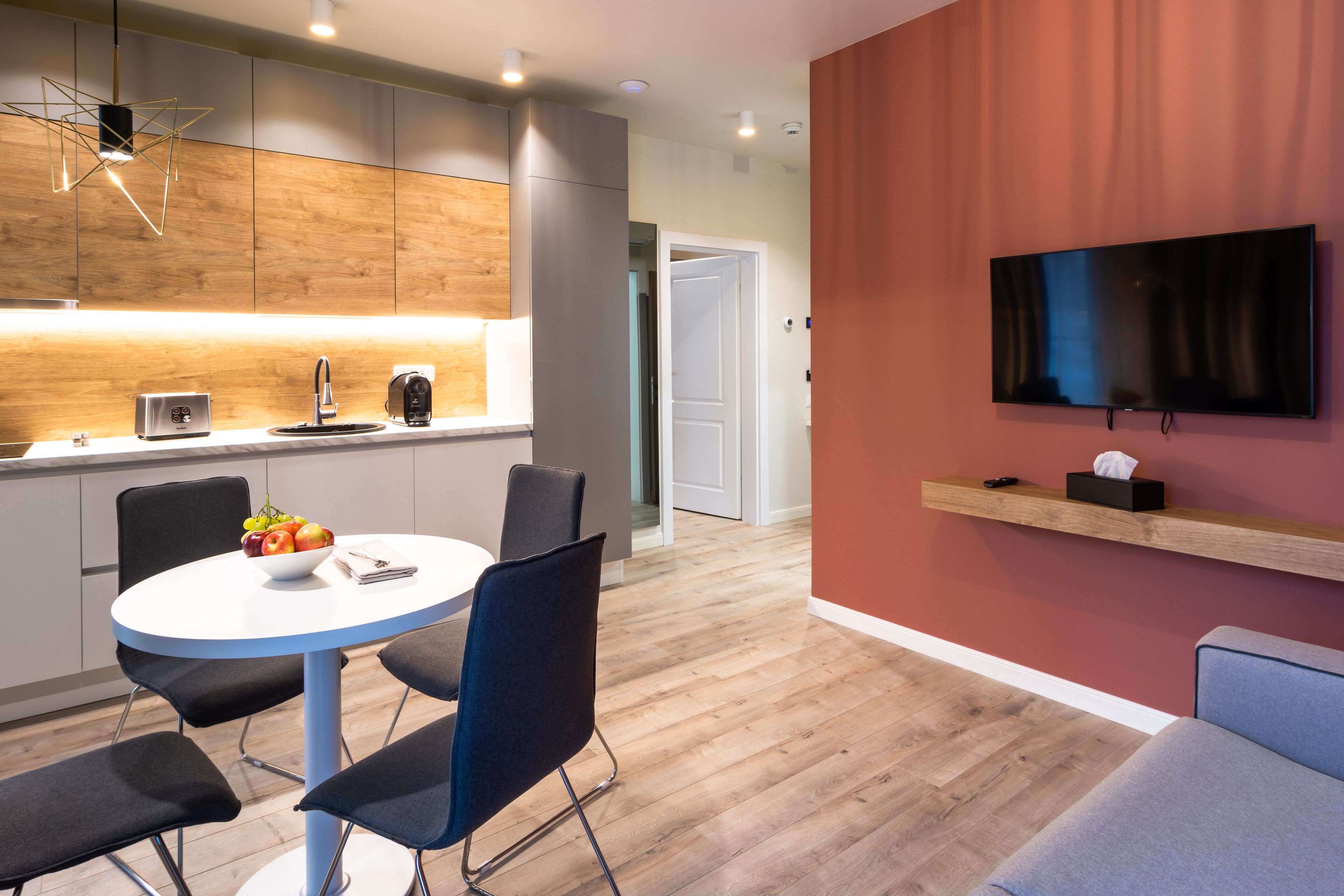 A modern kitchen with a dining area, a flat-screen TV mounted on the wall, and a wooden floor.