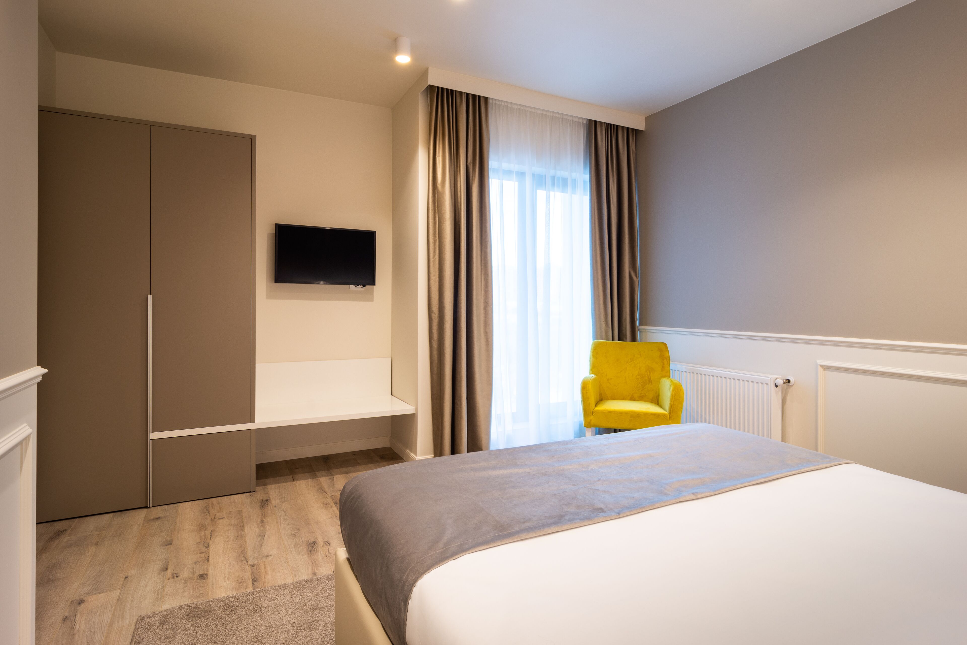 Una habitación de hotel con cama, televisión montada en la pared, una silla amarilla y un armario.
