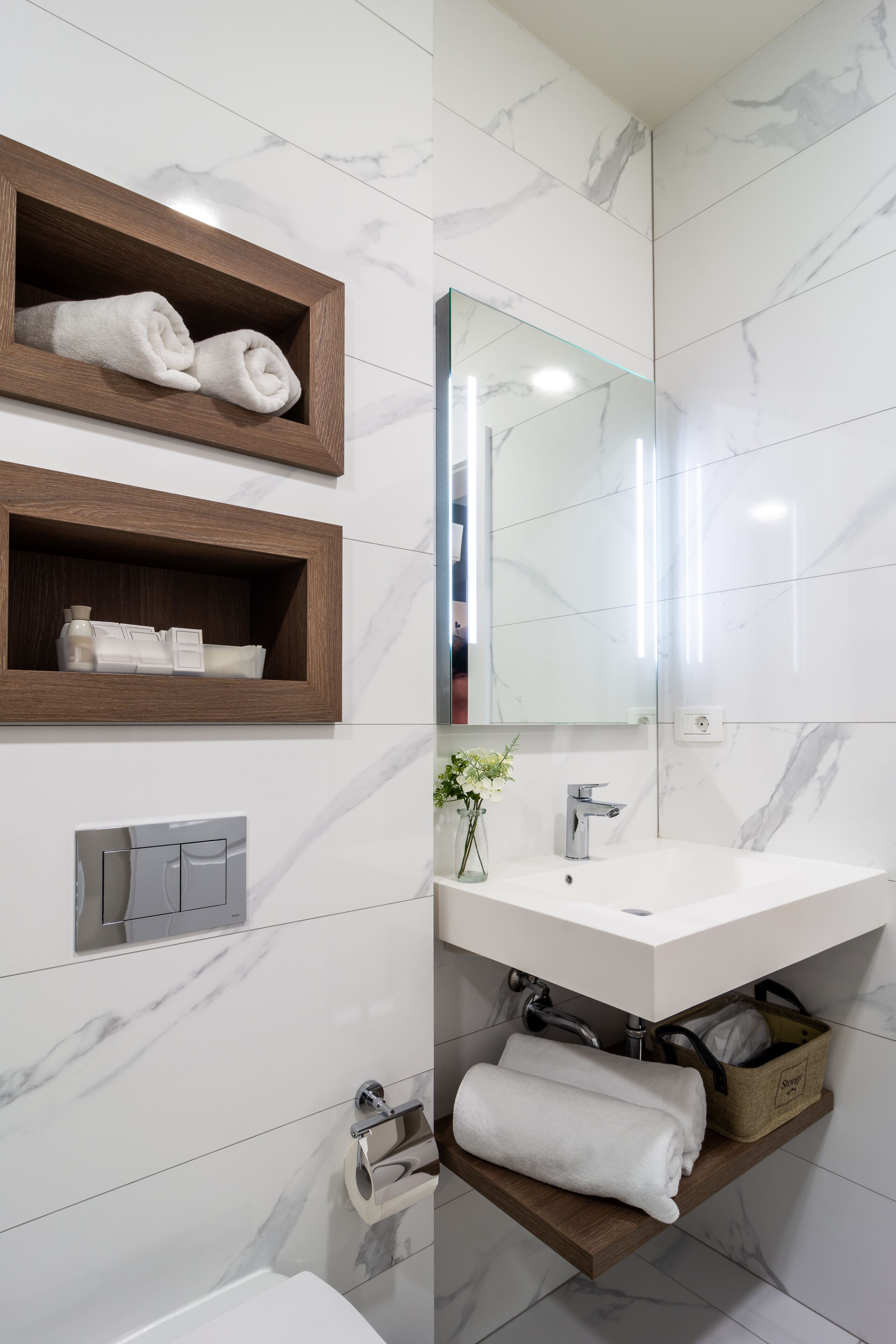 Une salle de bain moderne avec un pare-baignoire en marbre blanc, des étagères en bois, un grand miroir, un lavabo blanc et un porte-serviettes.