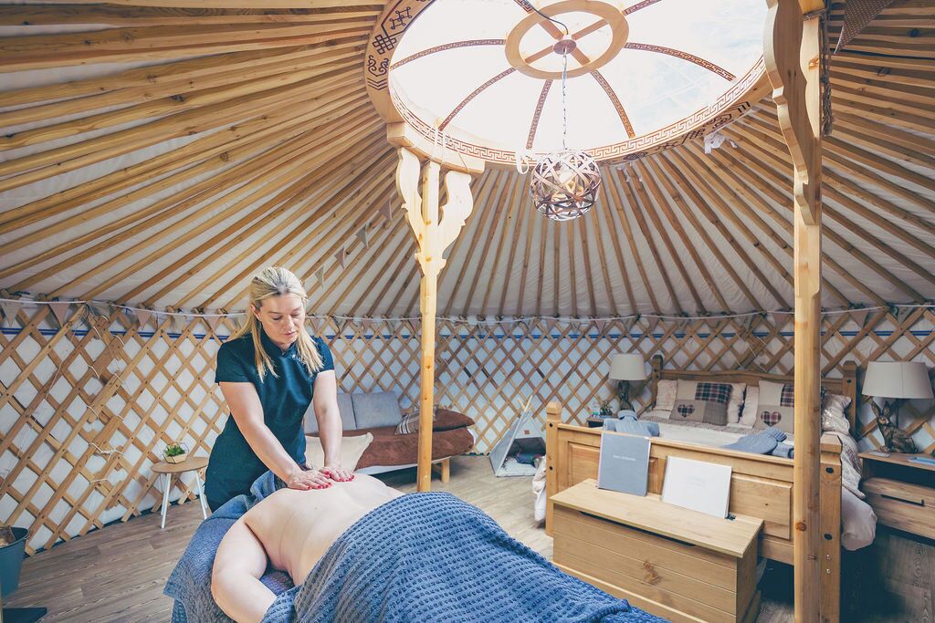 A yurt with a wooden interior, a bed, a massage table, and a massage therapist attending to a client.