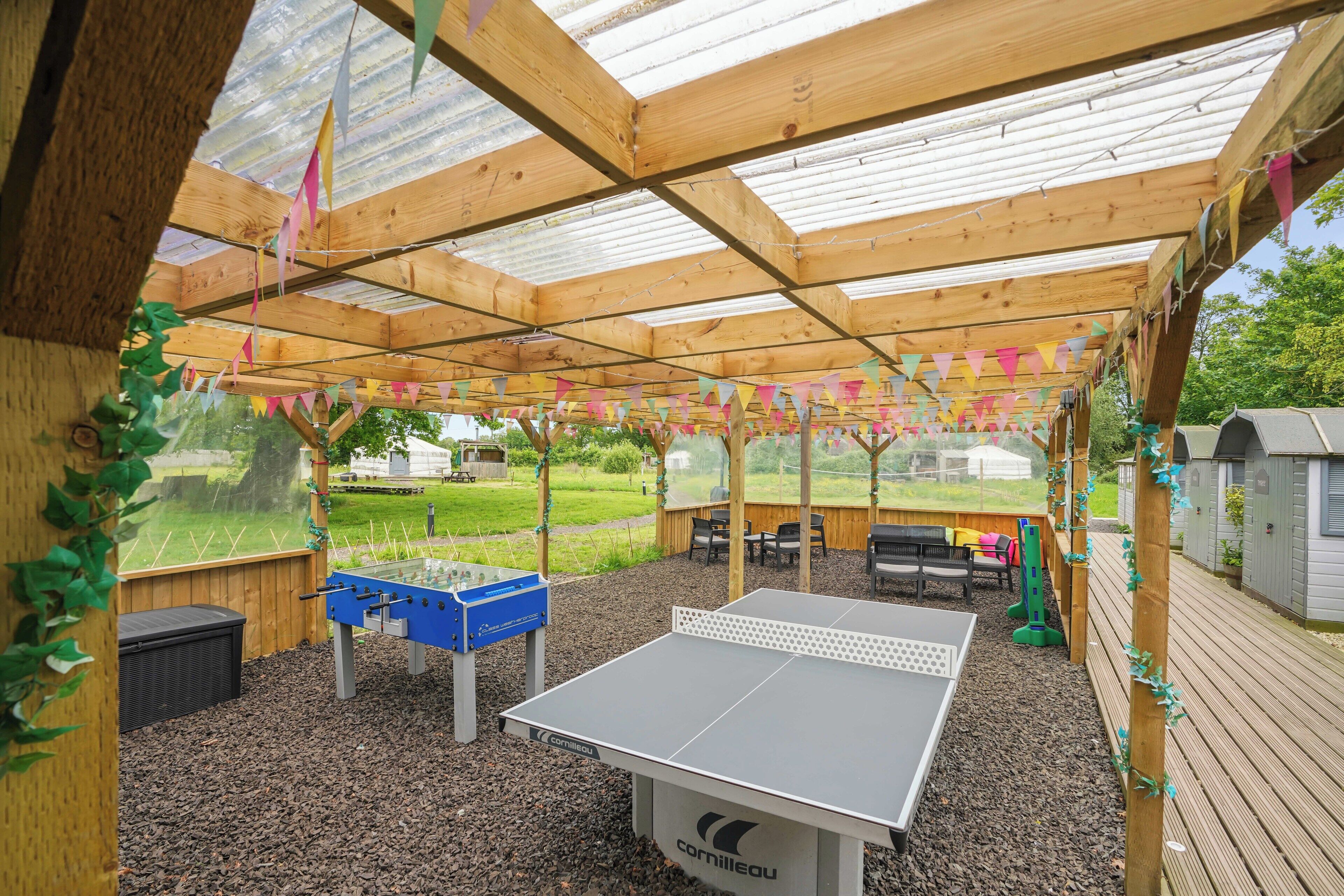 Een recreatiegebied buiten, met een tafeltennistafel, een tafelvoetbaltafel en een zithoek onder een houten pergola, versierd met vlaggen in verschillende kleuren.