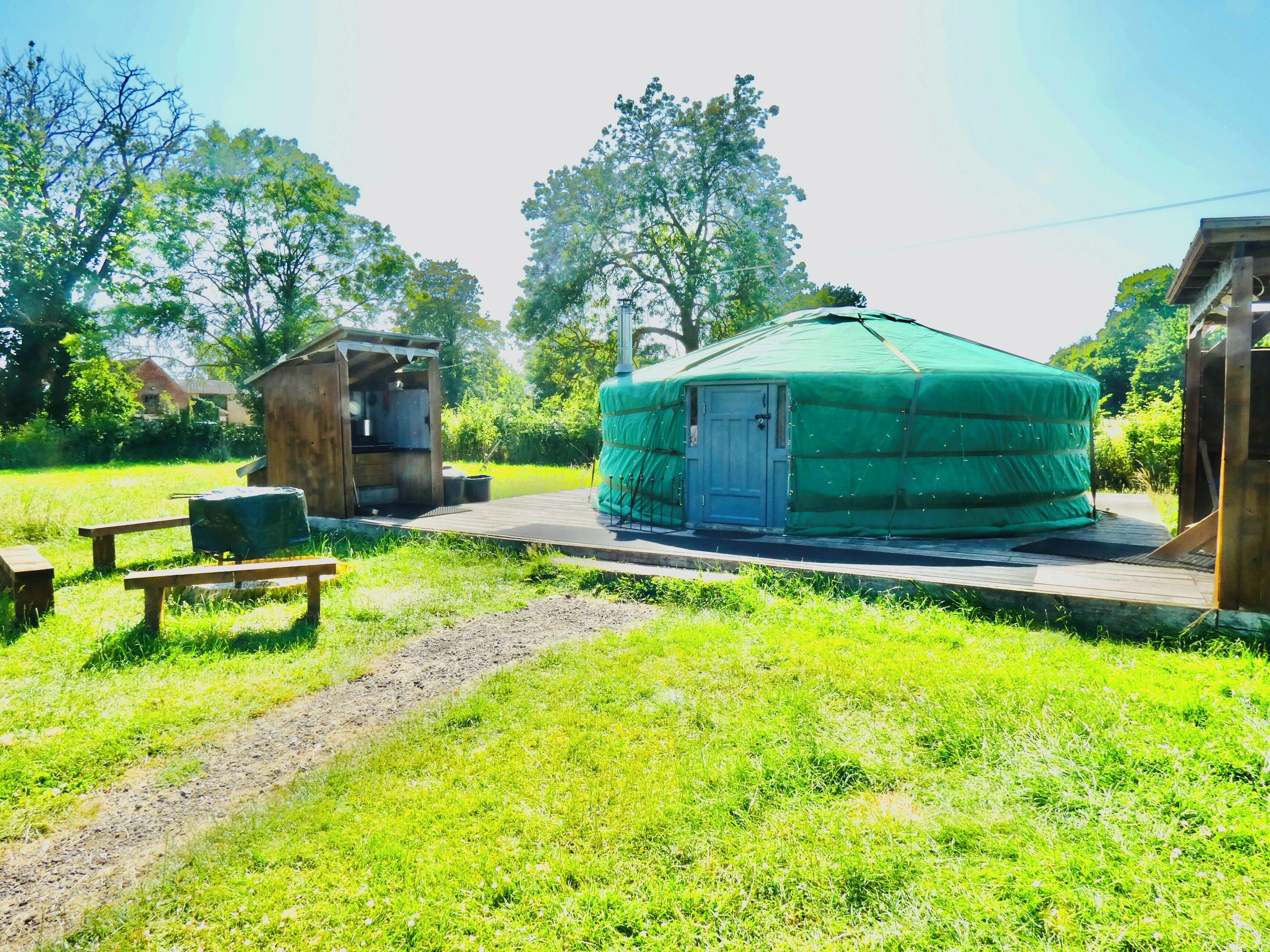 Een groene yurt met een deur, een klein houten constructie en een picknicktafel in een grasrijk gebied.