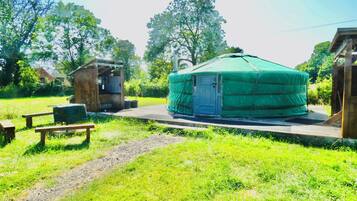 Een groene yurt met een deur, een klein houten constructie en een picknicktafel in een grasrijk gebied.