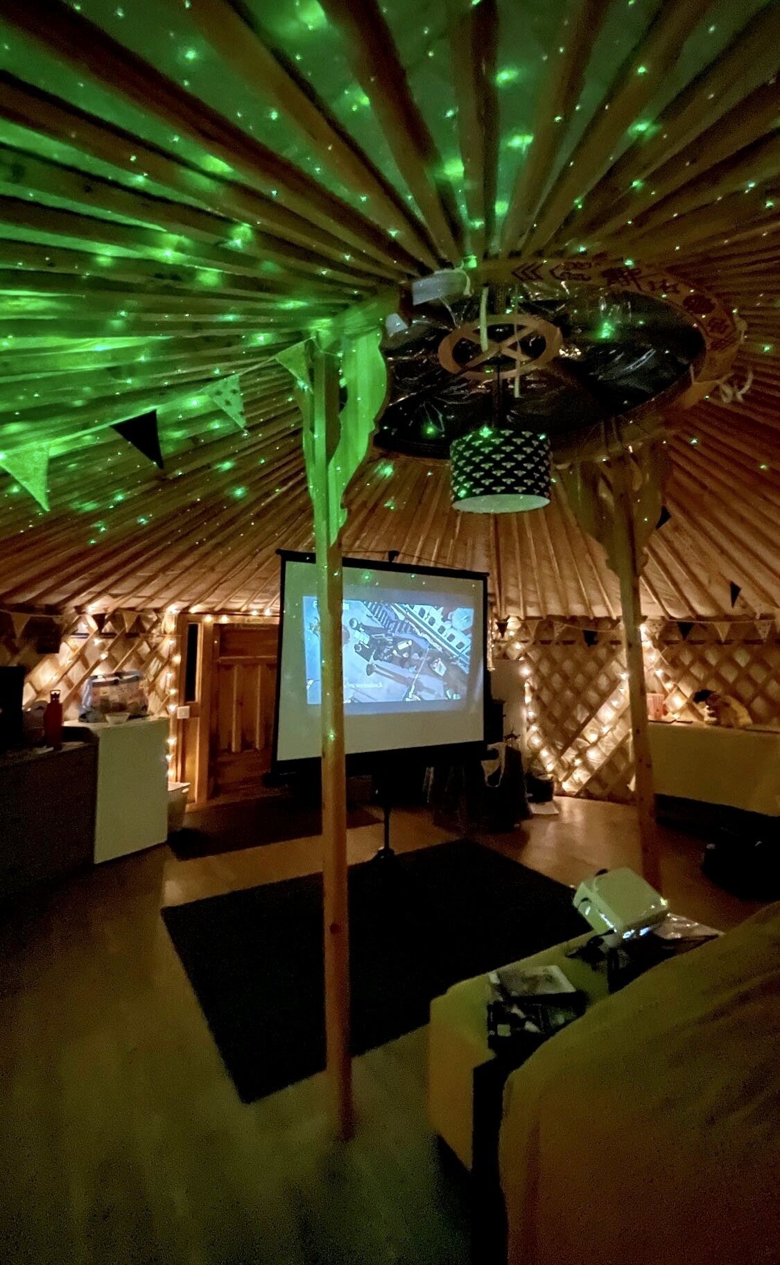 A cozy, dimly lit room with a large screen displaying a video, wooden furniture, and a unique ceiling design with green lighting.