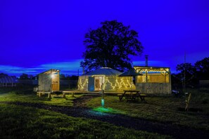 A rustic wooden cabin with a large tree in the background, a picnic table, and a small shed.