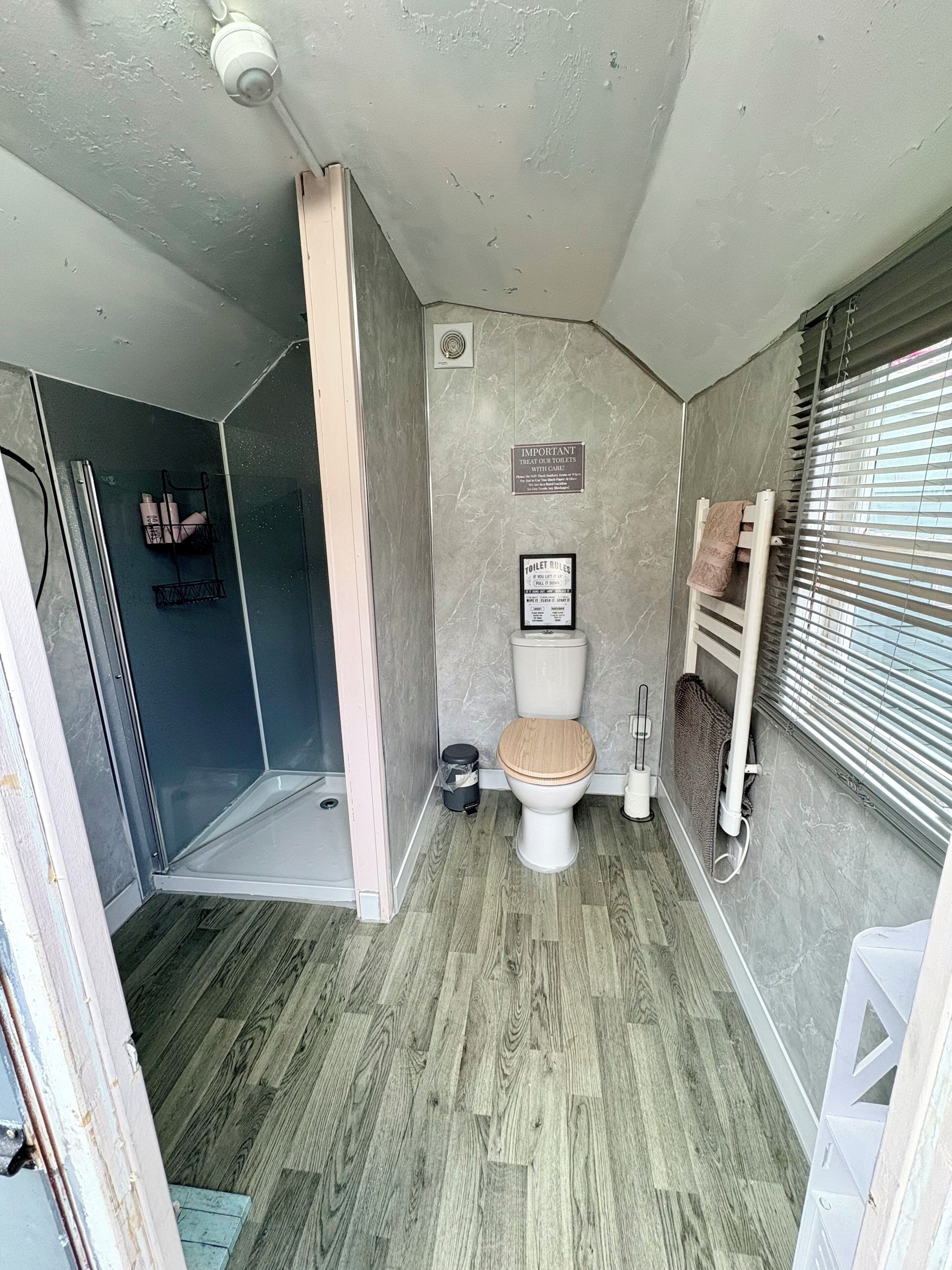 Une petite salle de bain comprenant une douche, des toilettes et une petite fenêtre avec des stores.