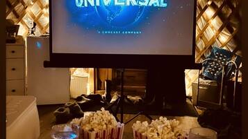 Una habitación acogedora con un televisor de pantalla plana grande mostrando el logotipo de Universal, recipientes de palomitas y dos vasos sobre una mesa de madera.