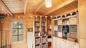 A wooden cabin interior with a wooden ceiling, a window, a bookshelf, storage bins, and a kitchenette area.