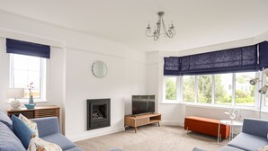 Una sala de estar con dos sillones azules, una pequeña mesa auxiliar de madera con una lámpara, un televisor sobre un mueble de madera, una chimenea y un ventanal con cortinas azules.
