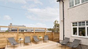 Una terraza de madera con cuatro sillas y una mesa, un edificio de fachada blanca y ventanas, y un cielo despejado.