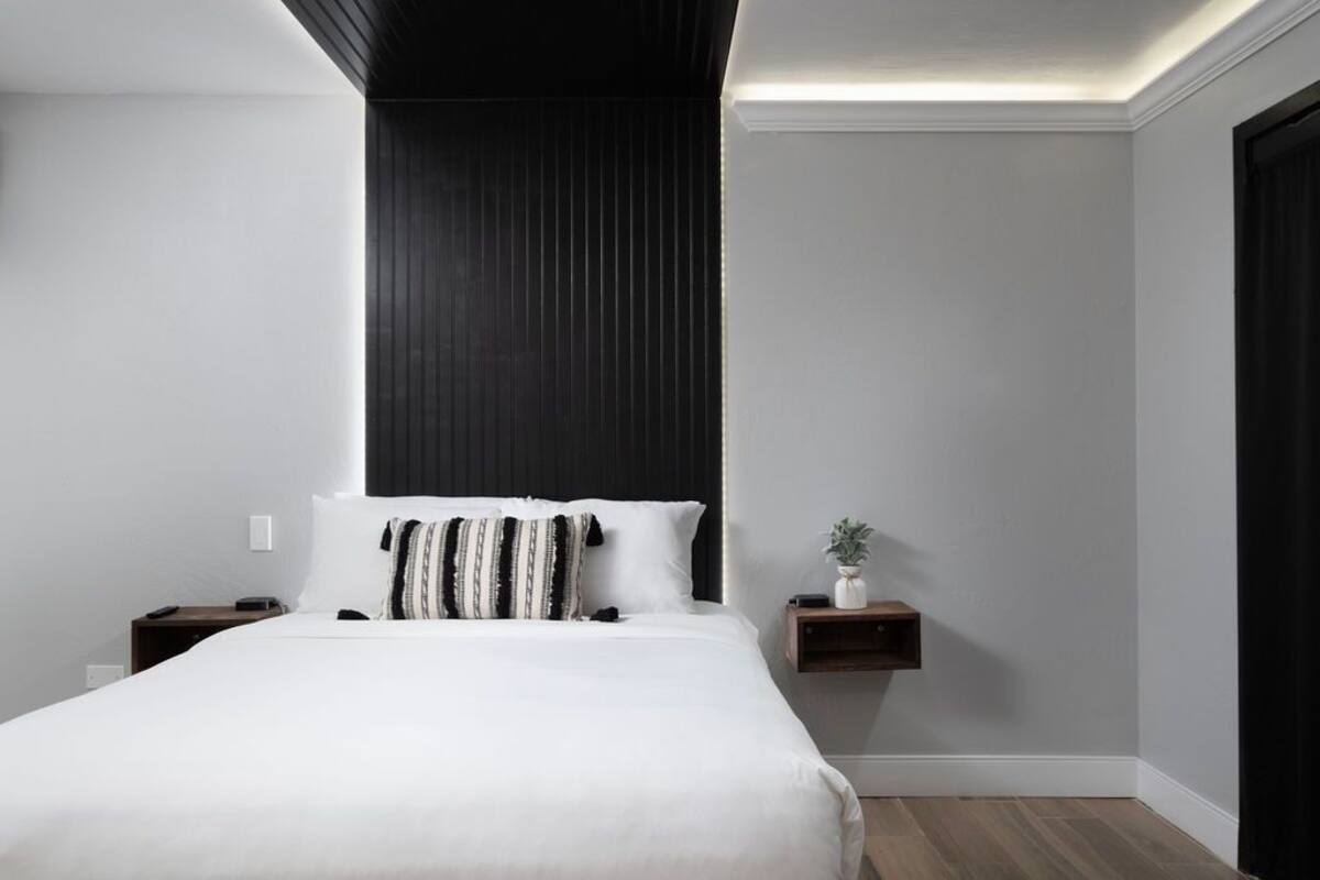 A modern bedroom with a large bed, a bedside table, a small shelf with a plant, and a dark accent wall.