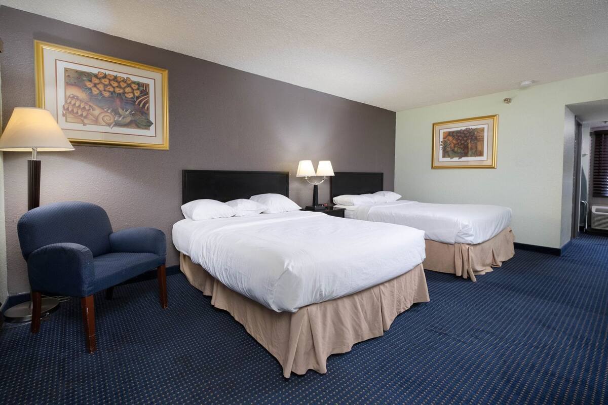 A hotel room with two beds, a chair, a lamp, and framed artwork on the walls.