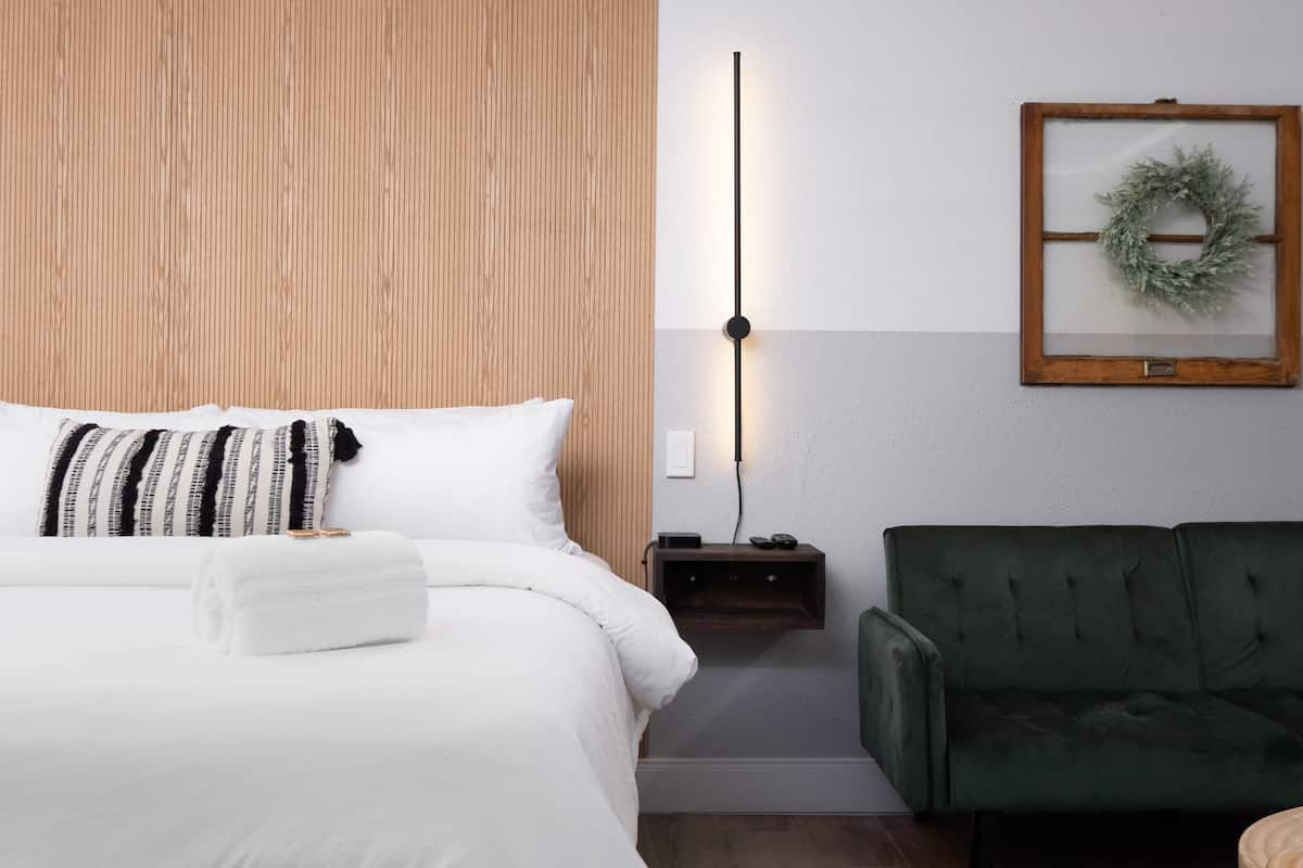 A neatly made bed with a white comforter and a folded white towel, a wooden headboard, a green upholstered sofa, a wall-mounted light, and a framed wreath on the wall.