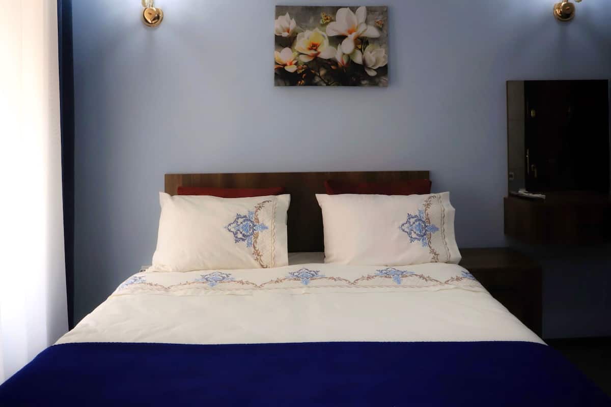 A neatly made bed with white linens and blue accents, a wooden headboard, and a floral painting on the wall.