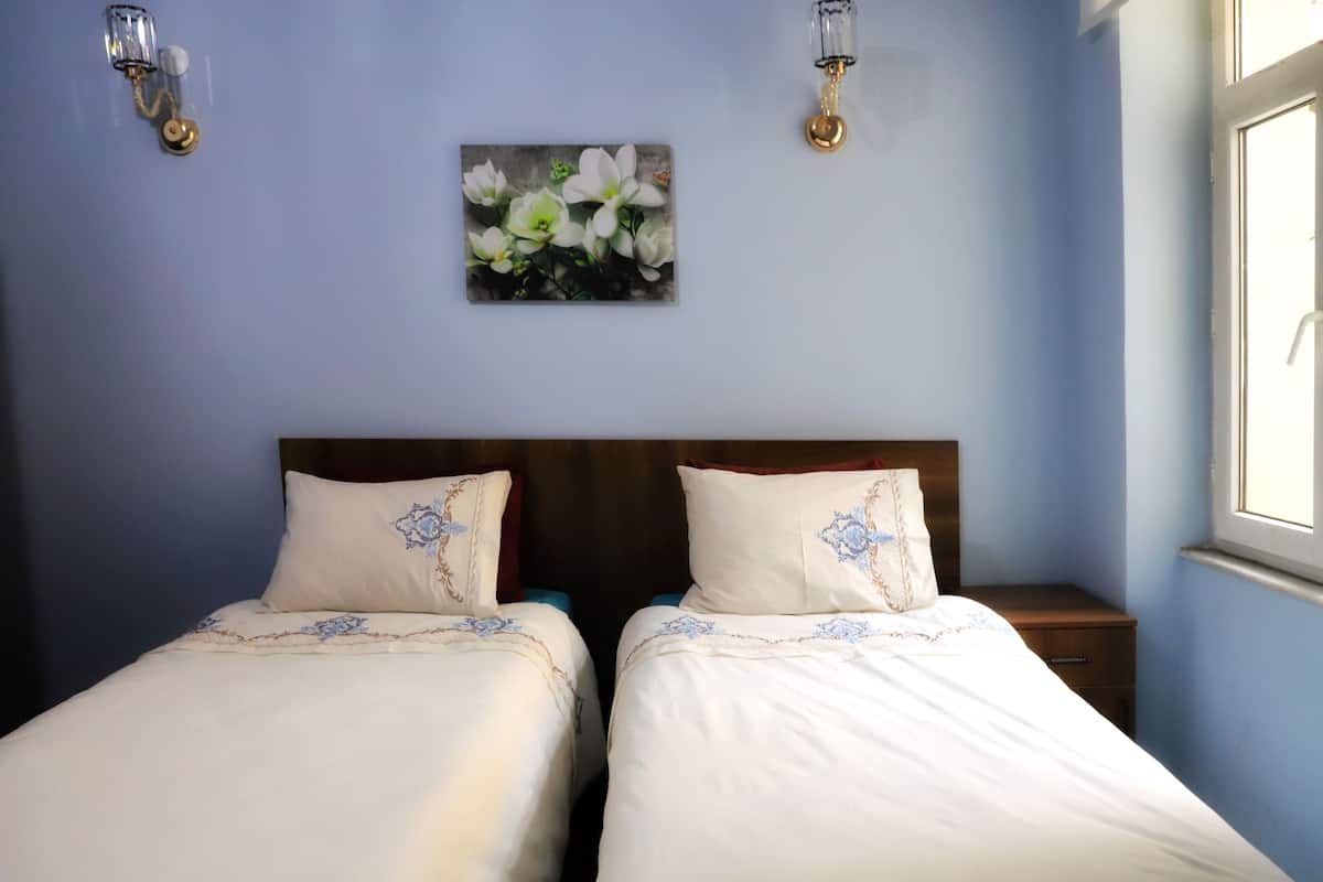 A bedroom with two beds, a nightstand, a wall-mounted light fixture, and a floral painting.
