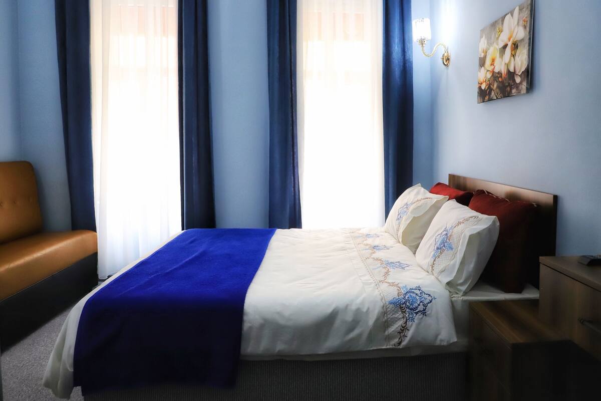 A hotel room with a bed, a nightstand, a lamp, a painting, and a window with curtains.
