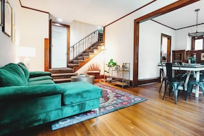 Ein Wohnzimmer mit einem grünen Sofa, einer Holztreppe, einem Essbereich mit Tisch und Stühlen sowie einer durch eine offene Tür einsehbaren Küche.