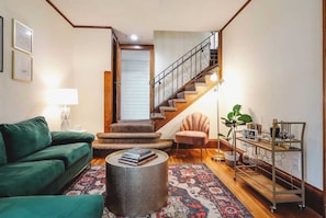 Una sala de estar con un sofá verde, una mesa redonda, una escalera, una silla y una estantería metálica con libros y vasos.