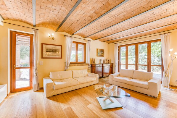 A spacious living room with two sofas, a glass coffee table, wooden flooring, and large windows with curtains.