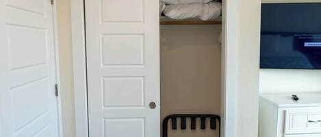 A room with a white door, a bed with white bedding, a radiator, a television on the wall, and a small cabinet with drawers.