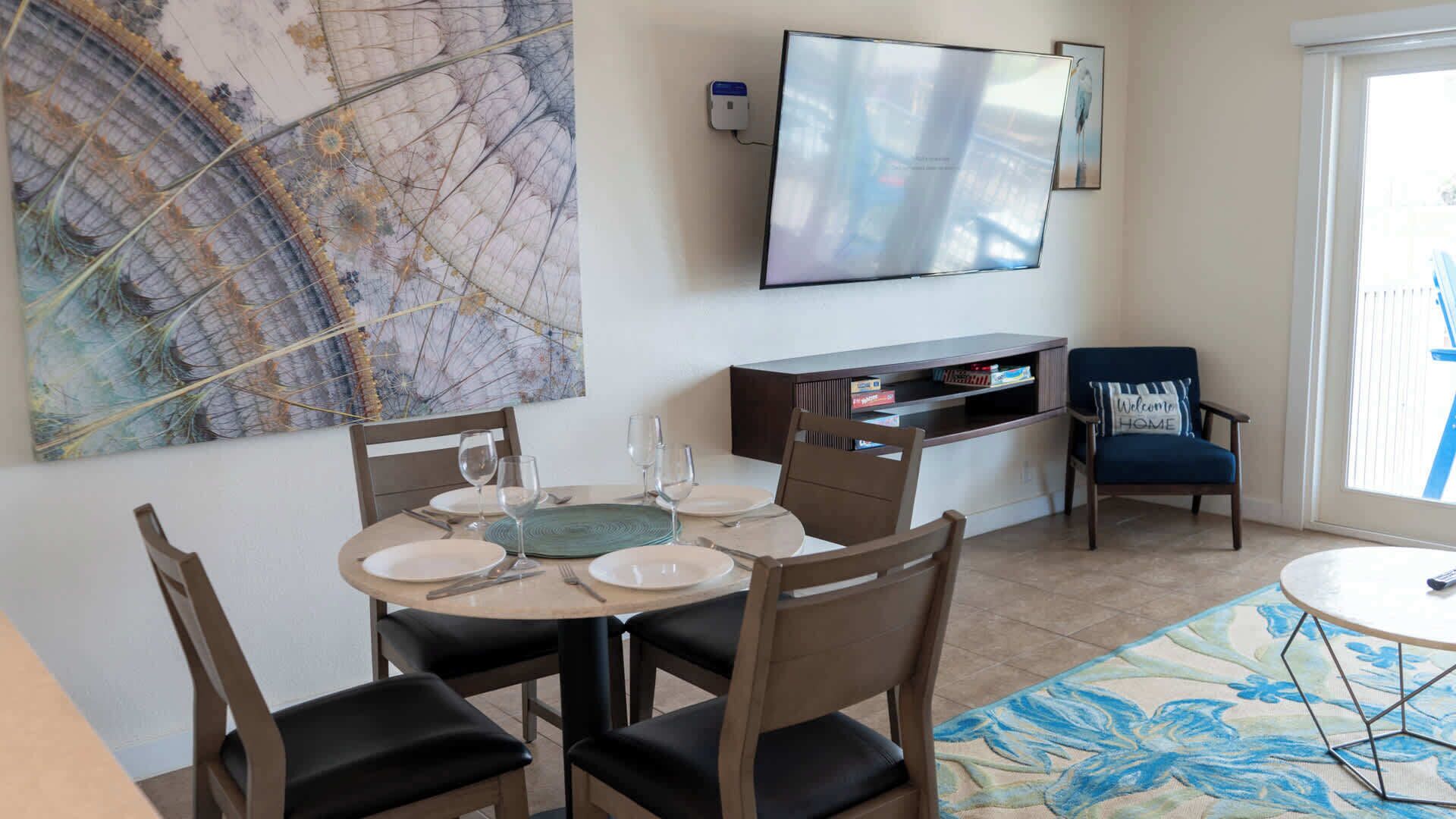 Un comedor con una mesa redonda dispuesta para cuatro personas, un televisor de pantalla plana montado en la pared y una estantería con libros y objetos decorativos.