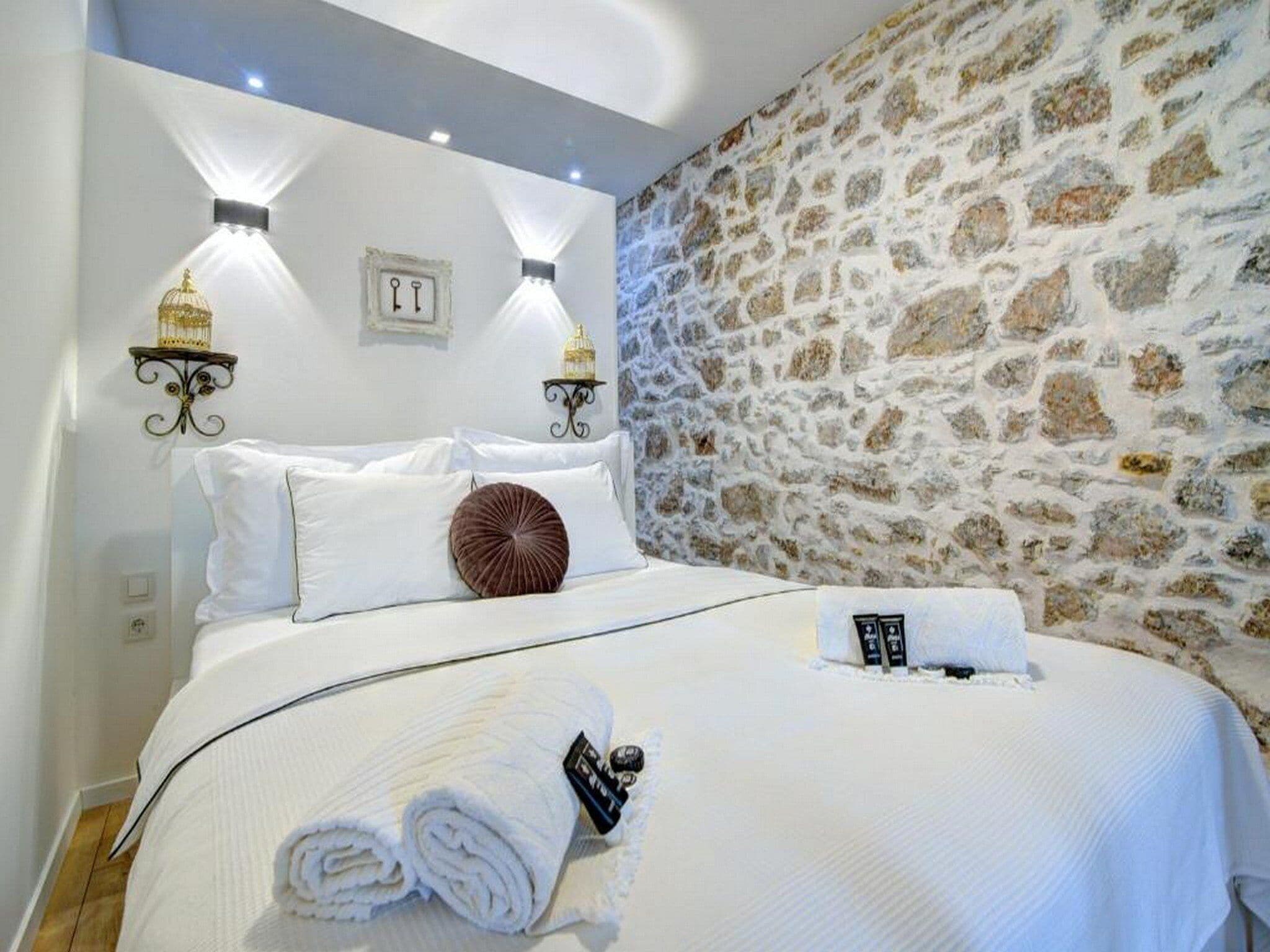 A neatly made bed with white linens, a rolled towel, and a remote control on the side. There are two wall-mounted light fixtures and a stone wall in the background.