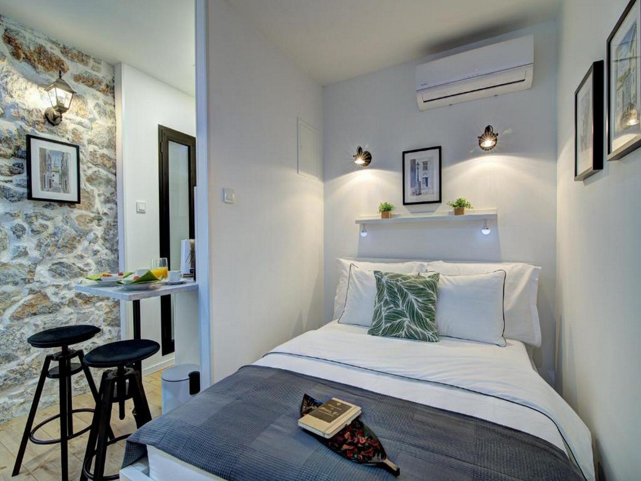 A bedroom with a bed, two bar stools, a small table with a chair, and a stone wall.