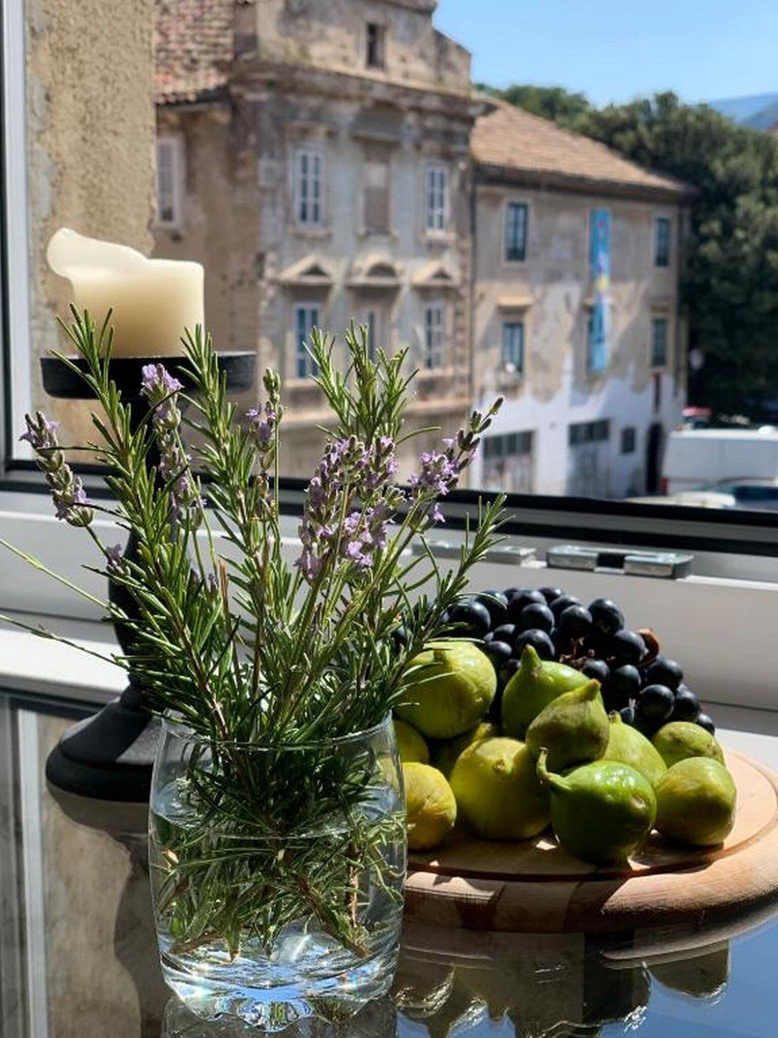A window with a vase of rosemary and a plate of fruits.