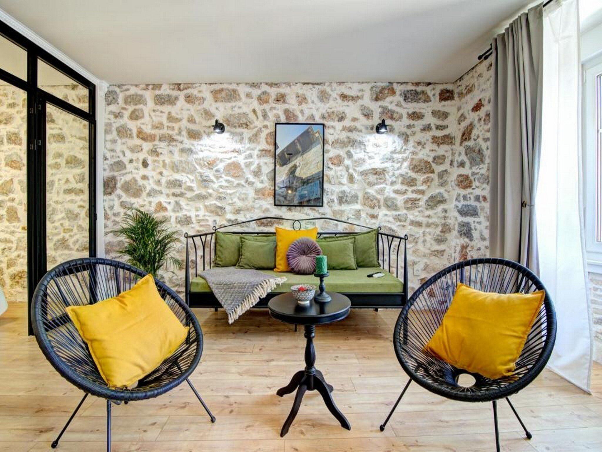 Um quarto aconchegante com parede de pedra, uma cama com roupa de cama verde, duas cadeiras de vime preto com almofadas amarelas e uma pequena mesa redonda com um vaso.