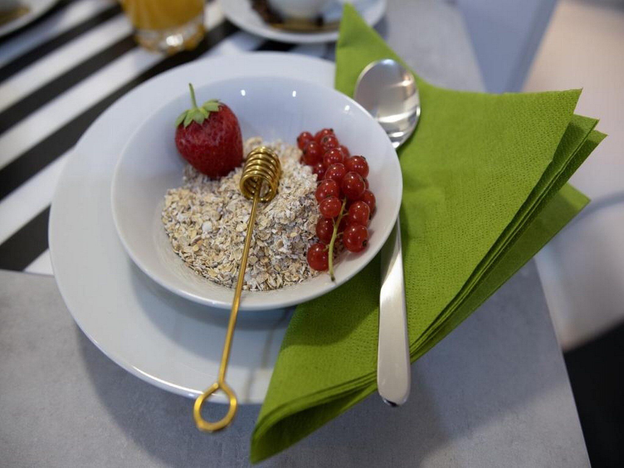 Colazione con una ciotola di porridge, fragole e ribes, insieme a un piccolo recipiente per il miele e un cucchiaio su un tovagliolo verde.