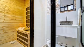 A sauna with wooden walls, a wooden bench, and a glass door leading to a bathroom with a stone sink and tiled floor.