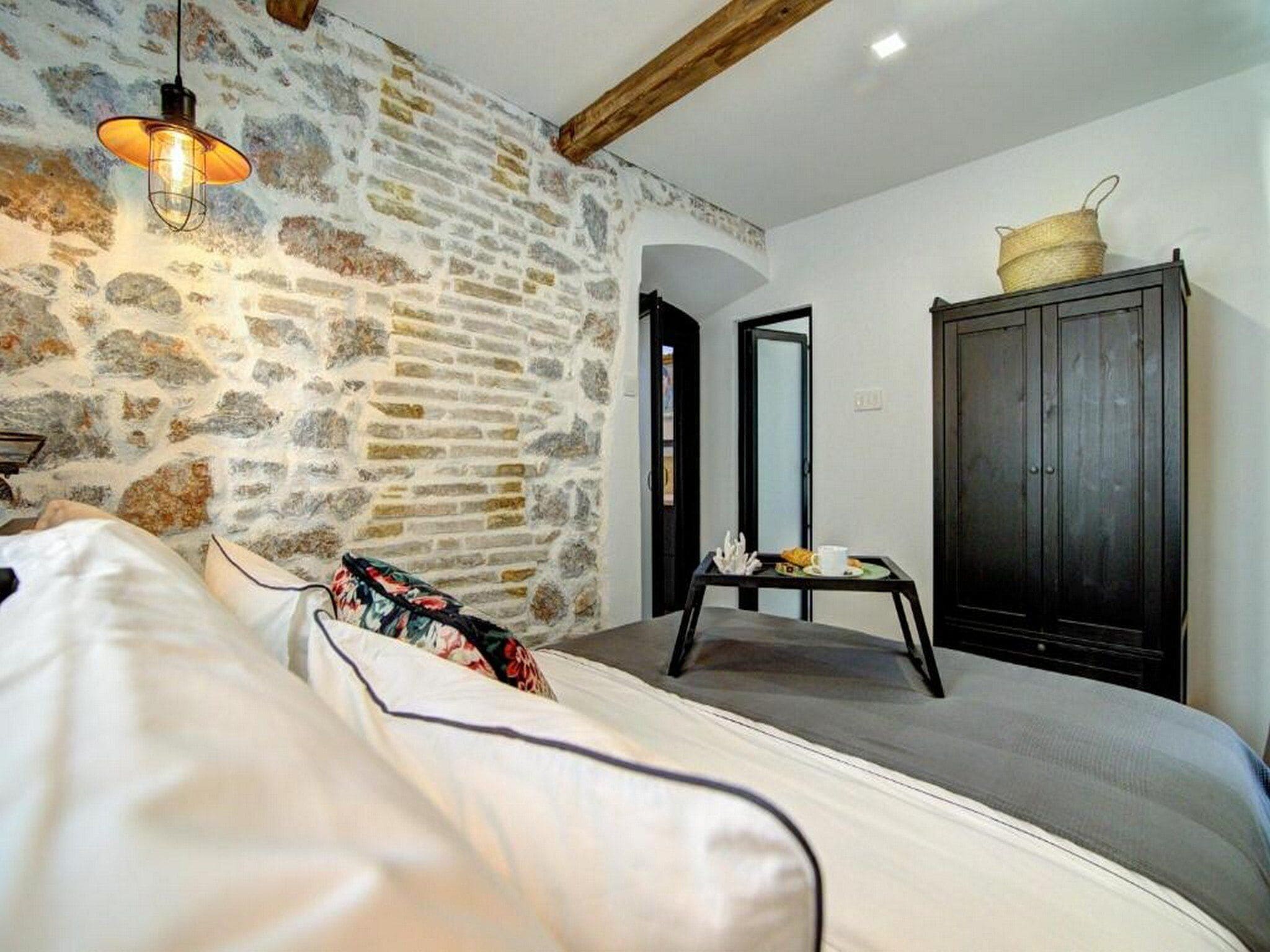 Un dormitori amb paret de pedra, llit amb roba de llit blanca, armari de fusta i taula petita amb una cistella a la part superior.