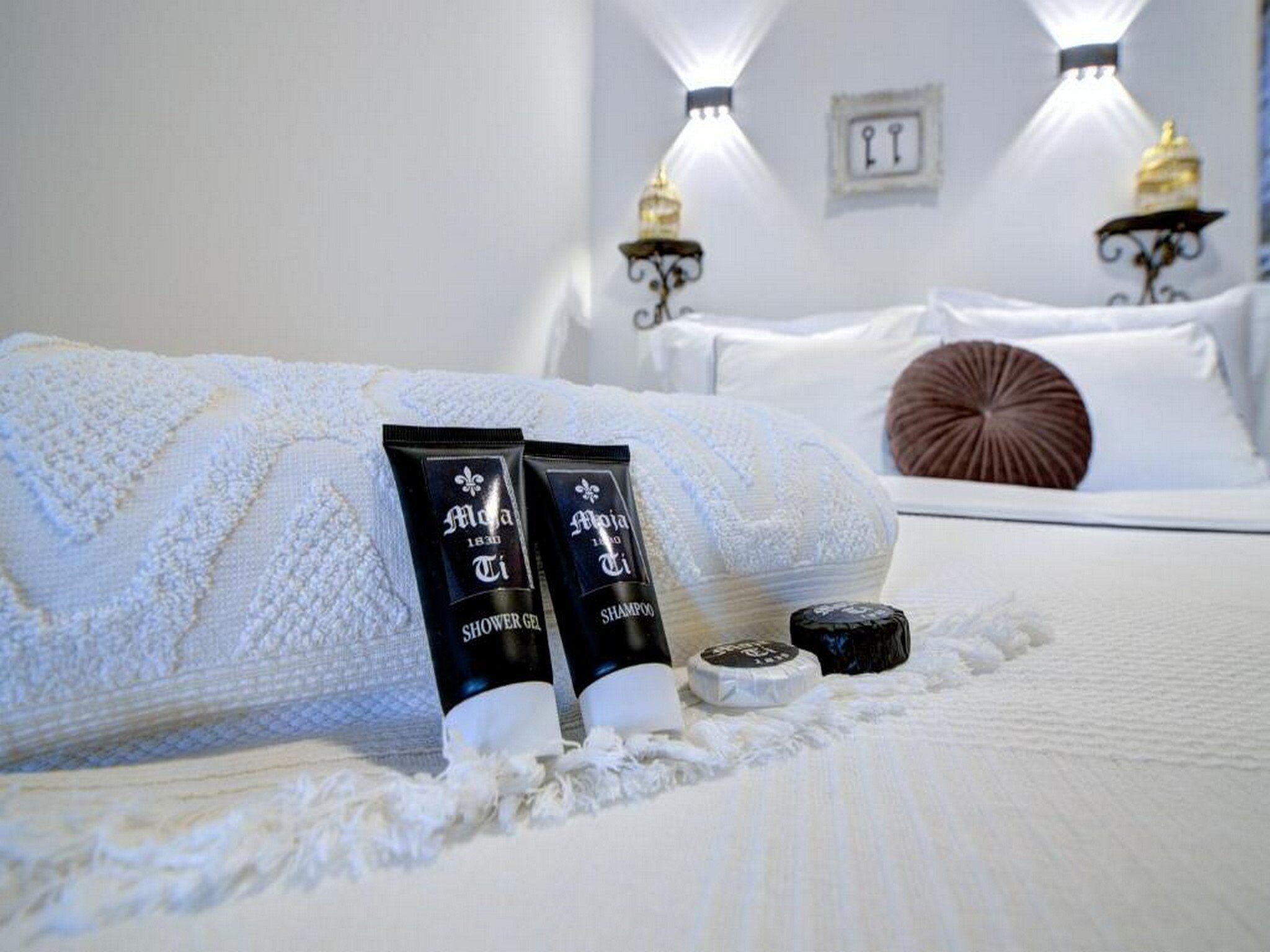 A neatly made bed with white linens, two black tubes of shower gel and shampoo, a round soap dish, and two wall-mounted lamps.