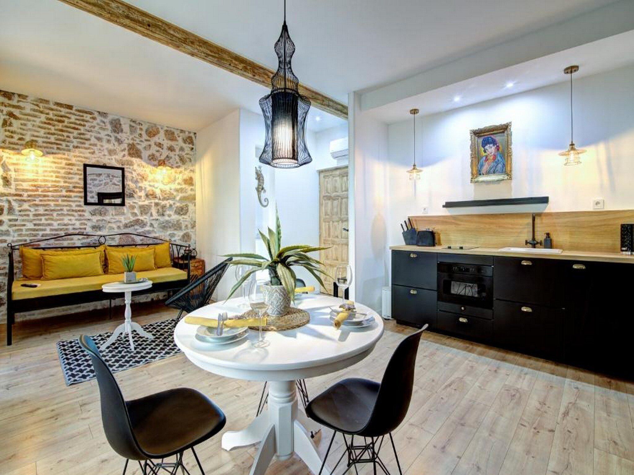 A modern living room with a stone wall, a sofa with yellow cushions, a round dining table with two chairs, and a kitchen area with black cabinets and a wooden countertop.