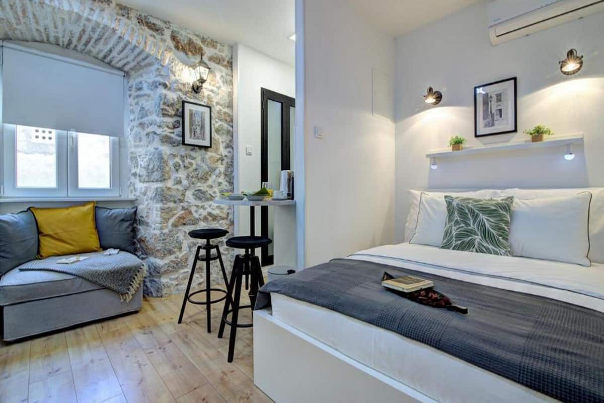 Un dormitorio moderno con una pared de acento de piedra, una cama con ropa de cama blanca, un sofá gris con cojines amarillos y una pequeña mesa con taburetes.