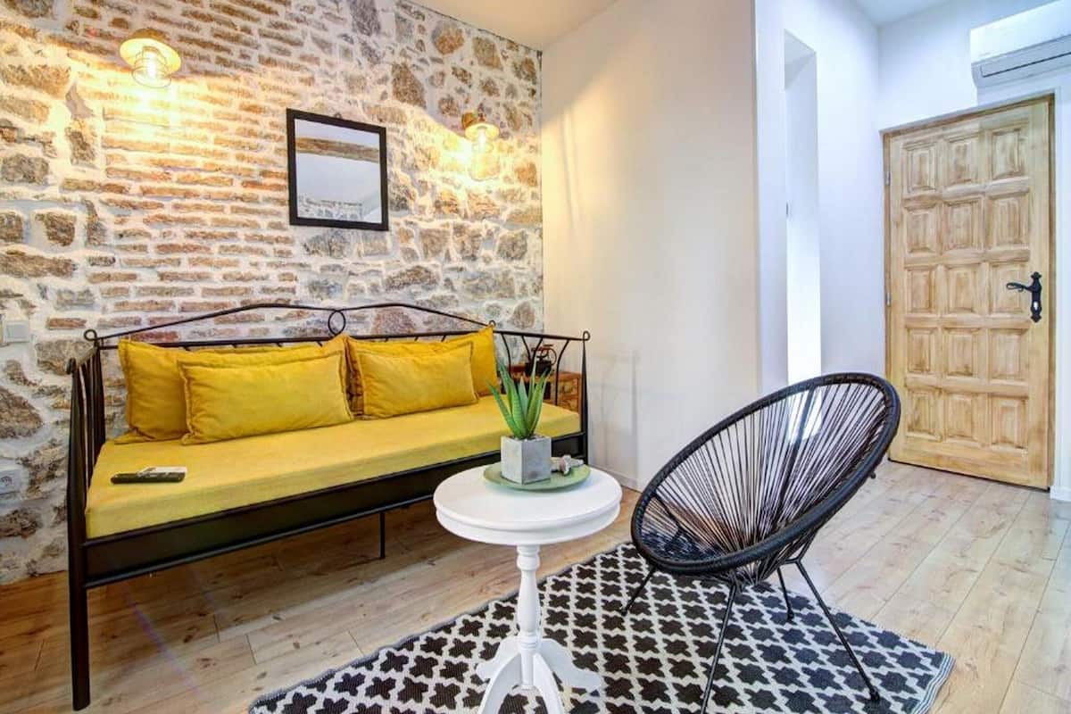 Una habitación con pared de piedra, un sofá amarillo, una mesa redonda blanca y una silla negra.