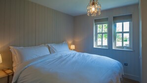 A bedroom with a large bed, two bedside lamps, a chandelier, and two windows with a view of greenery. - Remote & awe-inspiring Sealgair Cottage. Pet-friendly, log burner, lochside. (Shieldaig)