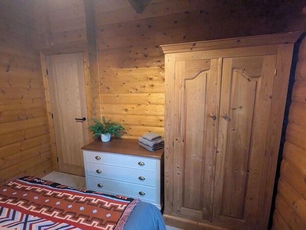 Una habitación de cabaña de madera con una cama, un aparador con cajones, un guardarropa y una planta en maceta sobre el aparador.