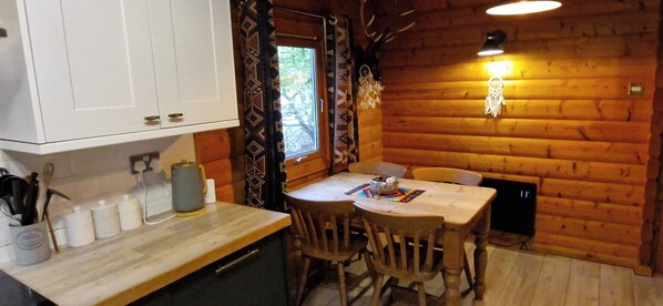 Un interior acogedor de cabaña con una mesa de comedor de madera, sillas, una zona de cocina con armarios y utensilios, y una cálida lámpara que crea ambiente.