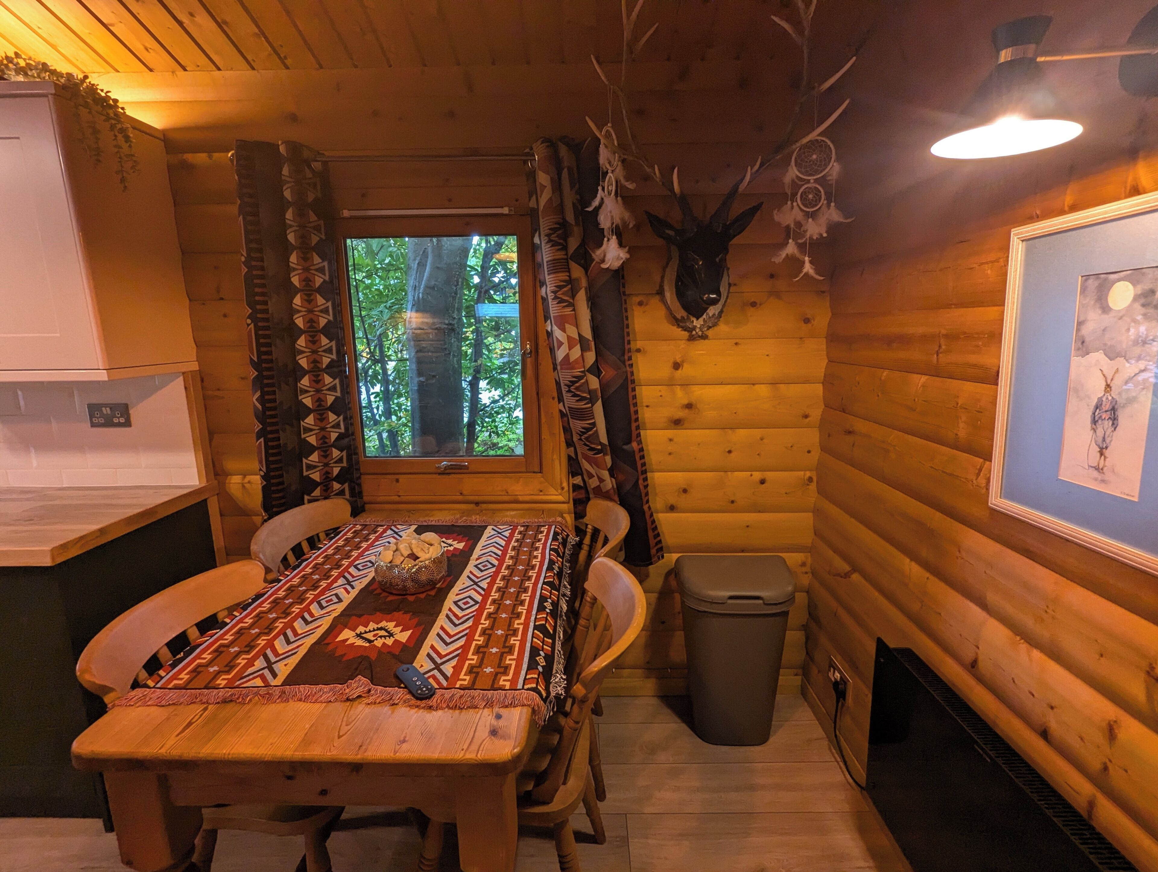 L’intérieur d’un chalet en bois comprend une table à manger, des chaises, une fenêtre avec des rideaux, une tête de cerf fixée au mur et un tableau encadré.