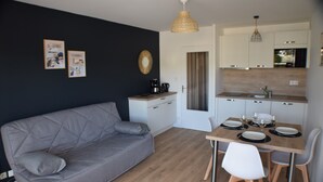 A modern living room with a grey sofa, a dining table with chairs, a kitchen area with white cabinets, and a hanging light fixture. - Ideal location for beach and pink granite coast. Sea view from the balcony (Perros-Guirec)