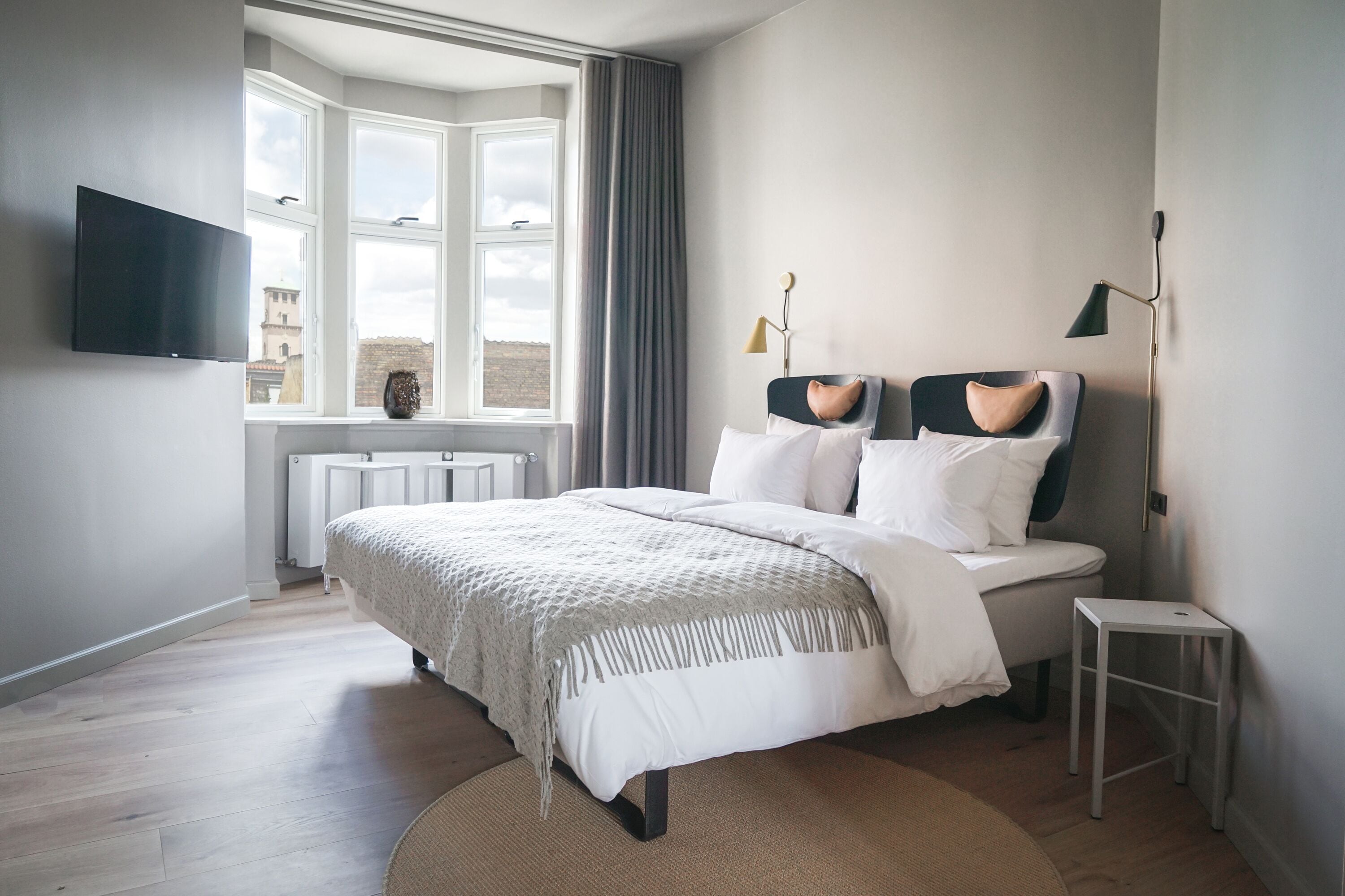 A modern bedroom with a large bed, a TV mounted on the wall, a window with curtains, and a radiator.