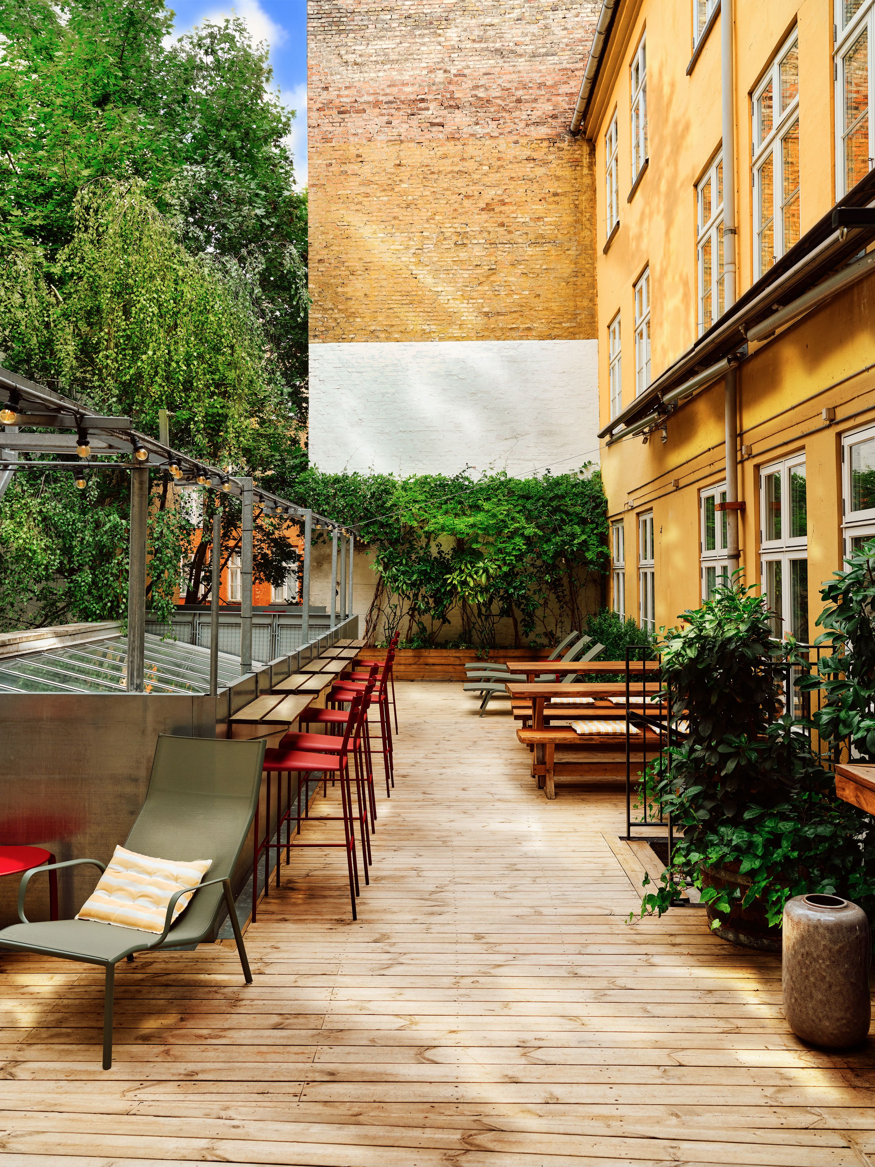 Eine Terrasse im Freien mit Holztischen und -stühlen, umgeben von Grünpflanzen und einer Backsteinmauer.
