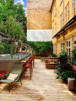 An outdoor patio with wooden tables and chairs, surrounded by greenery and a brick wall.