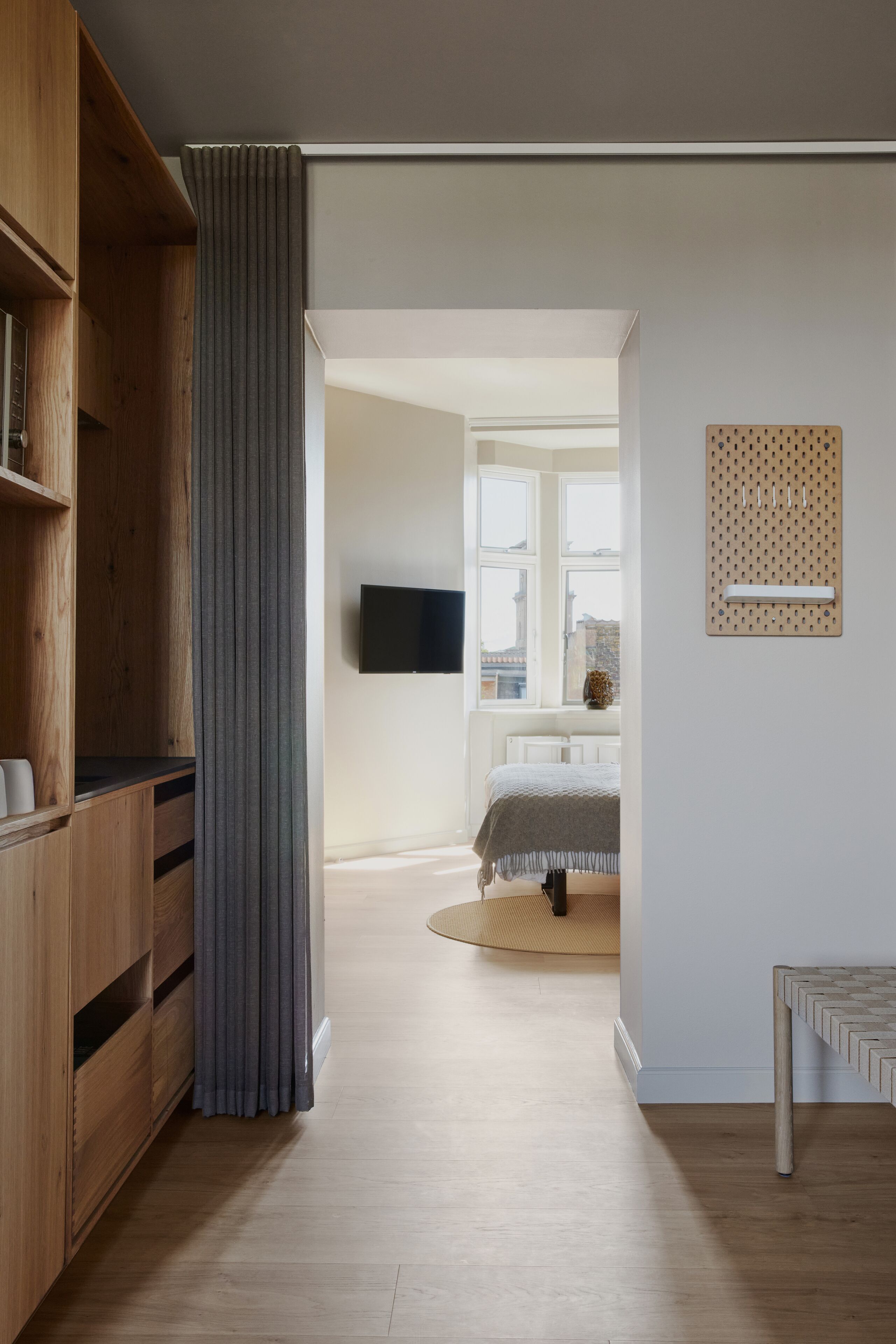 Une chambre moderne avec une armoire en bois, un rideau gris, un lit avec une tête de lit blanche, un téléviseur fixé au mur et une fenêtre donnant sur des immeubles.
